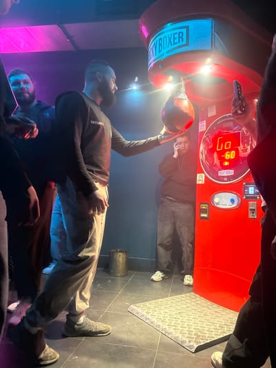 The image depicts an indoor scene, likely an entertainment venue, with a prominent red Rocky Boxer arcade machine. A man identified as security, wearing a black sweatshirt with "SECURITY" printed on it, is standing to the left of the machine, holding the punching bag. Other individuals are present, some partially obscured, suggesting a group setting. The machine has a digital display showing "-60". The environment appears dimly lit with colored lighting, creating a moody atmosphere. The flooring is tiled, and there is a metal mat in front of the machine. The overall context suggests a casual or recreational activity involving the boxing arcade game. There is no indication of the city of Kirov, Russia.