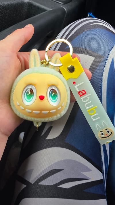 A close-up shot shows a person's hand holding a Labubu keychain. The keychain features a plush, light green creature with large, expressive eyes and a yellow face. It has small, white teeth-like protrusions around its mouth and a red heart-shaped nose. Attached to the plush toy is a light blue strap with the word "Labubu" written in raised letters. The "L" and "a" are in red, while the rest of the letters are white, with a yellow dot between the "L" and "a". A cartoon face with big eyes and a smile is also depicted on the strap. The background consists of a textured, dark material, likely part of a car interior, and a patterned fabric with blue, white, and gray stripes, possibly a car seat. The lighting suggests it is daytime, and the overall scene appears to be indoors, in a vehicle.