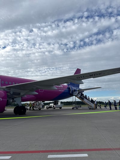 An Airbus A320-200 aircraft with registration 9H-WDK, operated by Wizz Air, is parked on the tarmac at Memmingen Airport (FMM) in Künersberg, Germany. The aircraft is predominantly pink and blue, with the Wizz Air logo visible. Passengers are boarding the aircraft via mobile stairs, indicated by the "ALLGATE" branding on the stairs. Several individuals, including passengers and ground crew, are present near the plane. The scene takes place under a cloudy sky with patches of blue. The ground is paved asphalt, marked with yellow and red lines. Trees and distant buildings can be seen in the background.