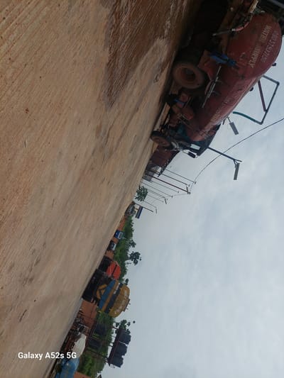 The image depicts a street scene in Awkuzu, Nigeria. The foreground is dominated by a textured, sandy-brown surface, possibly a road or pathway, with diagonal striations suggesting it has been raked or weathered. A portion of this surface appears wet and reflective.
In the mid-ground, several water tanker trucks are parked in a line. The closest and most prominent is a large, red tanker with "WATER SUPPLY" printed in white lettering and a phone number "08033793150". Another tanker behind it is blue and yellow with stripes and visible text that includes "NO SMOKING" and "HIGHLY INFLAMMABLE". Further down the line, other tankers in various colors can be seen, some with additional markings. The trucks are positioned at an angle, suggesting the photo was taken from a slightly elevated viewpoint or that the trucks are parked on a slope.
The background features a line of trees and vegetation, hinting at the presence of a green area or a more rural setting within the city. The sky above is overcast and a pale blue-grey. The overall impression is one of utility vehicles parked on a street, likely for a water distribution service. The image is oriented vertically, with the bottom of the frame appearing on the left. A watermark in the lower left corner reads "Galaxy A52s 5G".