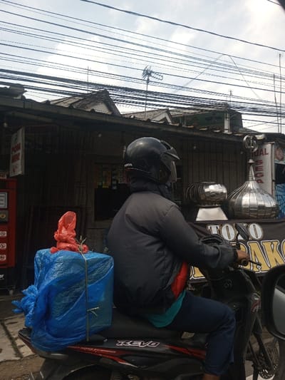The image depicts a street scene in an urban or semi-urban environment. While a location in the city of Chori, Nigeria was requested, prominent signage in the scene features Indonesian text, including "POM MINI" on a red self-service fuel dispenser, "TOKO" on a dark banner, and "JUAL" on a sign further right.
In the foreground, a single individual is seated on a black Honda Revo motorcycle, positioned facing right and slightly away from the camera. The person wears a black full-face helmet with a clear visor, a dark grey or black padded jacket, a teal shirt, and dark blue pants. The left hand is visible resting on the motorcycle's handlebar. Secured to the rear seat of the motorcycle is a large, rectangular package wrapped in bright blue plastic sheeting, fastened with green straps. On top of this blue package rests a smaller, irregularly shaped item wrapped in red plastic.
The background consists of a row of low-rise buildings, likely commercial or residential structures, topped with corrugated metal roofs. Various overhead electrical or communication cables crisscross the upper portion of the frame against an overcast, light grey sky. Multiple television antennas are visible on the rooftops. In the mid-ground, near the buildings, two shiny, metallic objects are present: one appears to be a turbine ventilator, and the other is a dome-shaped ornament, possibly a decorative finial or miniature mosque dome, displayed on a small platform. The individual and motorcycle appear to be stationary on a roadside or street.