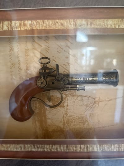 The image shows a framed decorative object. The object appears to be a replica of an antique flintlock pistol, made of metal and wood-like material. It is positioned horizontally, with the barrel pointing to the right. The pistol has intricate decorative carvings on the barrel. Behind the pistol, there is a background that resembles old parchment or paper with faded handwritten text. The frame has a wooden appearance with textured decorative bands. The lighting suggests an indoor setting, likely a room with natural or artificial light creating reflections on the glass of the frame. There is no information in the image that places it in Belo pole, Bulgaria or any specific city.