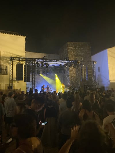 An outdoor nighttime concert is taking place in the historic center of Loulé, Portugal, as evidenced by the architecture. A band is performing on a stage erected with scaffolding and equipped with speakers and lighting. The stage is illuminated with bright yellow and blue lights, with beams of yellow light cutting through the darkness. A crowd of people is gathered in front of the stage, watching the performance. The crowd is densely packed, and many individuals are holding up their phones, likely to record the event. The background features traditional Loulé buildings, characterized by white-washed walls and stone elements, lit by the concert lights. The overall atmosphere is lively and festive.