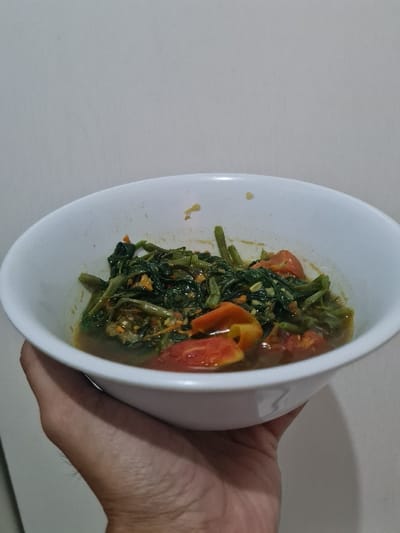 A white bowl held in a person's hand contains what appears to be cooked water spinach with tomatoes in a thin sauce. The spinach is mostly dark green, and the tomatoes are red and yellow. The bowl is positioned at an angle, showcasing the contents against a light-colored background. The person's hand is partially visible on the right side of the bowl.