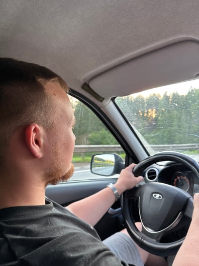 An eye-level, medium shot captures a person driving a vehicle on a road. The driver, a white male with short, light brown hair and a reddish-brown beard, is seen from the neck up, facing right and looking out the side window. He is wearing a black t-shirt and grey shorts. His left hand is on the steering wheel, a silver watch is visible on his left wrist. The interior of the car is visible, including the steering wheel, dashboard with an orange dial, and a sun visor. Outside the car, through the windshield and side window, a blurry green forest is visible, with sunlight filtering through the trees, suggesting it is either sunrise or sunset. A road barrier runs parallel to the car. The overall impression is that of a casual drive, possibly on the outskirts of a rural or suburban area. The location is specified as Opalikha, Russia.