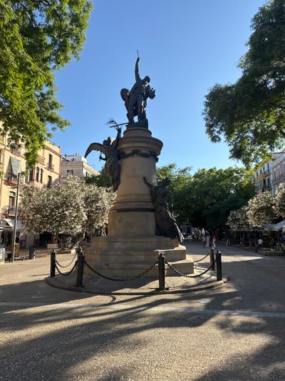 The image shows a monument in a public square in Eivissa, Spain. The monument is made of bronze and stone and features several figures, including what appears to be an angel with wings and a group of people. The monument is surrounded by trees, some of which are in bloom with white flowers. In the background, there are buildings with balconies and outdoor seating areas for cafes or restaurants. The ground in the square is paved with textured material, and shadows from the trees are cast across it. A chain fence surrounds the base of the monument. Several people are visible in the background, walking through the square. The sky is clear and blue, indicating a sunny day.