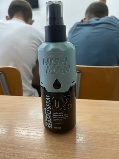 A close-up, eye-level, medium shot captures a bottle of "NISH MAN TEXTURIZING SEASALTS SPRAY" positioned centrally on a light-colored wooden desk. The bottle, primarily dark brown with a light green textured cap, features prominent white text. On the cap, the brand name "NISH MAN" is clearly visible, along with the text "CREATIVE MEN DESIGN." Below the cap, the bottle's label displays "TEXTURIZING SEASALTS SPRAY" vertically, followed by "02" in large numbers, and smaller text indicating "made with kelp & aloe vera," "marine rich texture spray," "nourishing," "texturizing," and "for all hair types," and "voluminous look." The volume "200mle" is also displayed.

In the background, out of focus, two individuals are seated facing away from the camera, suggesting they are students in a classroom or lecture hall. Their backs are to the viewer, with one wearing a white t-shirt and the other a blue shirt. The environment appears to be a classroom with wooden desks and chairs. The image lacks specific identifying landmarks to pinpoint its location within Svishtov, Bulgaria.