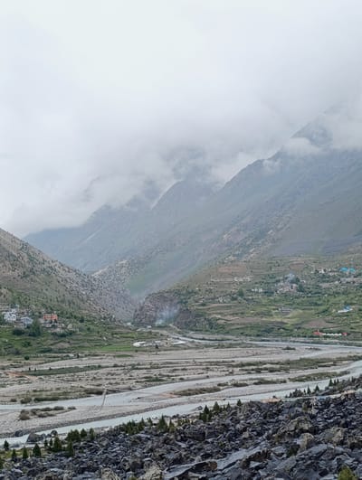The image captures a wide mountain valley in Darcha, India, under a heavy overcast sky. The foreground is dominated by a field of dark, jagged rocks, interspersed with a few sparse evergreen trees. Beyond the rocks, a wide, light-colored river or dry riverbed snakes through the valley floor, with smaller channels and gravelly banks.
Further into the valley, scattered settlements are visible on the lower slopes of the mountains. These villages consist of clusters of houses with predominantly white and colored roofs, built on terraced land. Some agricultural fields, appearing as green patches, are also present in these cultivated areas.
The mountains themselves are steep and rugged, with layered geological formations. The upper slopes are covered in dry, scrubby vegetation and bare rock, and the peaks are partially obscured by thick, grey clouds that hang low in the sky, creating a sense of depth and atmosphere. A faint plume of smoke is visible rising from a point within the valley, possibly from a dwelling or a small fire. The overall impression is one of a remote, rugged, and sparsely populated mountainous landscape.