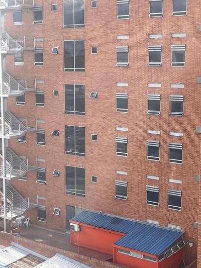 A high-angle exterior view captures a section of a multi-story building facade in Bogota, Capital District, Colombia. The facade is constructed from reddish-orange brick. On the left, a silver-grey metal fire escape with multiple levels of stairs and railings ascends vertically. The brick wall features a variety of window types: several large, dark-paned rectangular windows are present on the left and central portions, interspaced with smaller square windows. To the right, numerous rows and columns of uniform rectangular windows are visible, each equipped with grey, horizontal exterior roller blinds; many of these windows are tilted inward from the top, indicating a partial opening. Below the main facade, a flat rooftop terrace extends. Situated on this terrace is a long, rectangular, reddish-orange structure with a blue corrugated metal roof and an external air conditioning unit mounted on its side. Adjacent to it is a smaller, L-shaped structure, also with a blue corrugated metal roof and windows. Near the base of the fire escape on the terrace, a small white plastic table with two white plastic chairs is present. In the foreground, at the bottom of the image, the weathered corrugated metal roofs of adjacent lower structures are partially visible. No individuals are depicted in the scene.