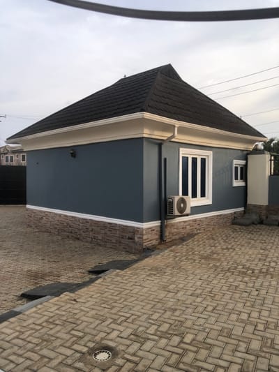The image displays a single-story residential building situated within a paved compound in Egan, Nigeria, under an overcast sky. The structure features a dark brown, tiled hip roof with white fascia and gutters. Its exterior walls consist of a muted blue-grey upper section and a lower section clad with horizontal, multi-toned brown and grey stones, separated by white trim. On the visible right facade, there are two white-framed windows with dark vertical panes, and an outdoor air conditioning unit is mounted beneath the larger window. A white downspout runs vertically along the corner. A small, dark exterior light fixture is mounted on the left-facing wall. The ground is paved with light-colored, rectangular interlocking concrete pavers arranged in a herringbone pattern. A dark drainage channel runs across the paved area, and a circular drain cover is visible in the foreground. Distant buildings are partially visible to the left, and overhead power lines traverse the top of the frame. No people or active interactions are depicted.