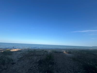 This media file depicts a serene beach scene at Bliznatsi, Bulgaria, under a clear, bright blue sky. The image is dominated by the vast expanse of the sea, which stretches out to meet the horizon. Gentle waves lap at the sandy shore, creating a peaceful and tranquil atmosphere.
The foreground is composed of sand dunes covered with sparse, dry grass and other low-lying vegetation. There is a noticeable shadow cast on the sand, suggesting the sun is positioned high in the sky, likely indicating midday or early afternoon. The overall lighting is bright, with the sun illuminating the water and creating dappled patterns on the sand.
In the middle ground, near the water's edge, a piece of driftwood is visible partially submerged in the water. The sea itself appears calm with a slight ripple effect on its surface. The distant horizon shows a faint hazy line, hinting at land or coastline. A very small, faint crescent moon is visible in the upper right quadrant of the sky, indicating it is daytime.
The scene evokes a sense of calm and natural beauty. There are no people or animals visible in the frame, emphasizing the quiet solitude of the location. The clear sky and calm sea suggest pleasant weather conditions, ideal for a day at the beach. The image appears to be a landscape photograph taken to capture the essence of this coastal location.