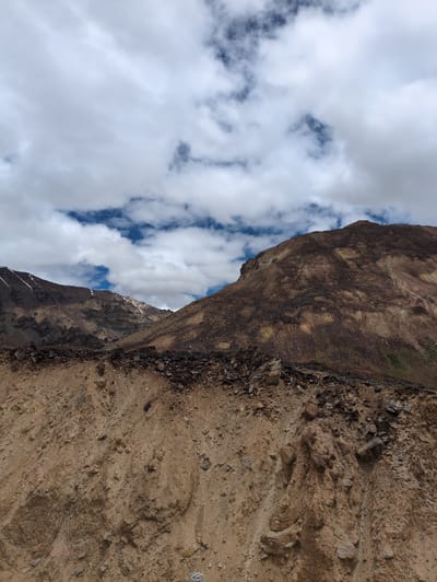 The image shows a mountainous landscape with a cloudy sky. In the foreground, there is a steep, rocky slope with brown soil and scattered stones. The midground features more rugged, brown-colored mountains with visible layers of rock and some patches of snow near the peaks. The background is dominated by a sky filled with dramatic white and gray clouds, with patches of blue sky visible in between. There are no people or man-made structures visible in the image. The scene appears to be in a natural, arid, or semi-arid mountainous region. The location is identified as Killingsarai, India.