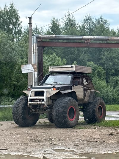 This media file shows a custom-built monster truck with extremely large tires. The truck is dark in color and appears to be heavily modified for off-road use, featuring a reinforced bumper and what looks like a toolbox or storage compartment on the back. It is parked outdoors on a dirt or gravel surface, with some mud or water visible. In the background, there are lush green trees and a cloudy sky, suggesting it might be a temperate climate.

The truck is the primary subject, and no people are visible. The setting appears to be a rural or possibly an industrial area given the large pipes running vertically and horizontally, with some of these pipes appearing weathered and rusted. The overall impression is one of ruggedness and power, suitable for extreme terrain. The lighting suggests it is daytime, but the overcast sky indicates potentially cloudy weather. The image is oriented vertically, with the truck appearing to be on its side. There are no discernible signs with visible text. The scene suggests a location in Yayva, Russia, possibly a recreational area for off-road vehicles or a maintenance yard.