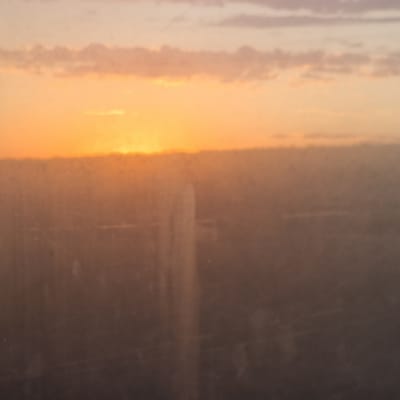 The image depicts a hazy, out-of-focus sunset over a low-lying, indistinct landscape, presumably the city of Badam, Kazakhstan. The sky is filled with soft hues of orange, yellow, and pale purple, with layered clouds contributing to the diffused light. The lower half of the image is dominated by a blurry, brownish-grey expanse, which could represent urban structures obscured by atmospheric conditions or distance. A vertical, lighter-toned blur is visible in the mid-left portion of this area, possibly a building or a significant landmark. The overall impression is one of a muted, almost surreal twilight scene, lacking sharp details and clear subjects.