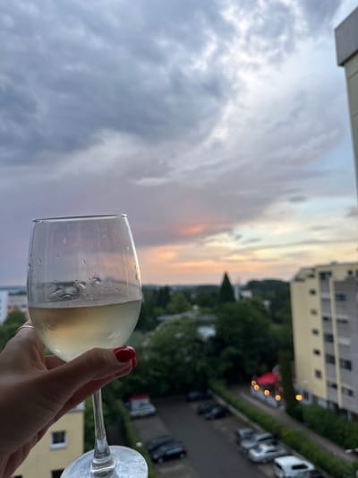 A hand with red nail polish holds a stemmed glass of white wine in the foreground, obscuring a view of a city street. The glass is partially filled with a pale yellow liquid and condensation. In the background, a dramatic sky with gray and purple clouds is visible, with hints of pink and orange near the horizon, suggesting sunset. Beyond the clouds, a cityscape with buildings and trees is visible. Cars are parked on a street below, and an apartment building is prominent on the right side of the frame. The scene is likely from an apartment balcony in Sindelfingen, Germany, overlooking a residential area.