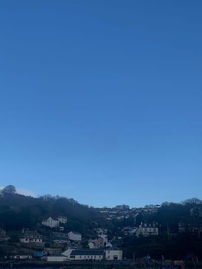 The image displays a panoramic view of the town of Looe, United Kingdom, situated on a steep, tree-covered hillside. Approximately the upper two-thirds of the frame are occupied by a clear, bright blue sky with no visible clouds. The lower third of the image features a densely wooded slope, predominantly with deciduous trees in a dormant state, indicating winter or early spring.

Numerous residential structures, primarily white or light-colored with dark roofs, are integrated into the contours of the hillside, appearing stacked from the base to near the crest. In the immediate foreground, at the very bottom of the frame, a waterfront or embankment area is visible. On the left, a multi-story building with dark windows is present. Towards the right foreground, colorful playground equipment, including slides and climbing frames, is discernible. Several dark, indistinct human figures are visible on the ground near this playground area. A horizontal barrier or fence runs along the bottom edge of the scene. The overall lighting suggests daylight conditions.