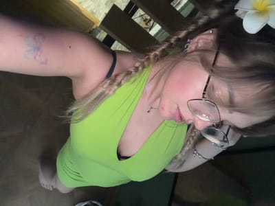 A young adult female, with blonde hair in braids and wearing a neon green halter dress and glasses, is standing indoors on a wooden floor. She has a tattoo on her left upper arm and is adorned with earrings and bracelets. A white flower is positioned in her hair. The background appears to be a staircase with wooden railings. The scene is not identifiable as being in the city of Большая Шильна, Russia.
