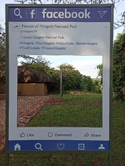The image displays a large, outdoor sign designed to resemble a Facebook post, serving as a photo frame. The sign is oriented vertically but the photo is taken with a 90-degree counter-clockwise rotation. Through the central cutout of the sign, a natural outdoor scene is visible, featuring bright green grass, several trees with dense foliage, and a reddish-brown pathway leading towards a small building or hut partially obscured by trees. The sky above is clear, indicating daytime.

The sign is bordered in a dark blue color, mimicking Facebook's interface. At the top left corner, the Facebook logo and a search icon are visible. The "user profile" section of the mock post reads "Friends of Akagera National Park" with the handle "@Akagera NP". The main text of the "post" states: "I visited Akagera National Park." followed by a series of hashtags: "#Akagera", "#OurAkagera", "#AfricaParks", "#IamInAkagera", "#VisitRwanda", and "#IVisitedAkagera". The bottom section of the sign features icons typical of Facebook posts, including "Like," "Comment," and "Share," alongside a home icon, a magnifying glass, a heart icon, and a plus sign. No people are visible in the frame. The sign clearly identifies its location context as Akagera National Park, in Mwiri, Rwanda, encouraging visitors to share their experience on social media.