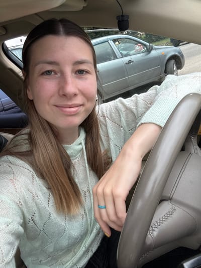 A young woman with brown hair, wearing a light green knitted sweater, is taking a selfie inside a car. She is in the driver's seat, with her left hand resting on the steering wheel. A turquoise ring is visible on her left ring finger. In the background, through the car window, another car is visible on a paved road, suggesting an urban or suburban setting. The overall scene is outdoors.