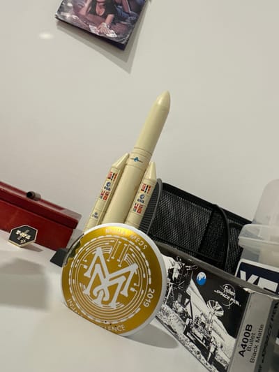 The image depicts a collection of objects arranged on a white surface, possibly a desk or shelf. The objects include a model rocket, a red box, a hexagonal pin, a circular sticker or badge, and a box for a pen. The model rocket is cream-colored with several stages and features logos that appear to be "esa" and national flags. The red box is rectangular with a metallic clasp. The hexagonal pin is black with a silver design and the letters "AS9". The circular badge is gold with a stylized "M" and text that reads "MUHA MEDS" and "INITIAL EXCELLENCE 2019". The pen box is black with a detailed illustration of a lunar landing and text that includes "Fisher SPACE PEN" and "A400B Bullet Black Matte". The background shows a white wall, a black mesh organizer, and a partially visible plastic container. There is no indication of the city of Zibatá, Mexico, or any specific environmental context. The scene is static and there are no people or actions taking place.