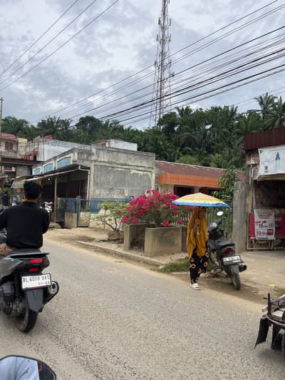The image depicts a street scene in Rantau Pauh, Indonesia. In the foreground, a person on a scooter is viewed from behind, riding on a paved road. To the right, a woman wearing a yellow hijab and patterned pants holds a colorful umbrella over her head. She stands next to a parked motorcycle with license plate BL 5384 UJ. The background shows a row of buildings, some with faded paint, and lush green trees on a hillside. A tall telecommunications tower is visible in the distance, along with a cloudy sky. Commercial signs are present on some of the buildings.