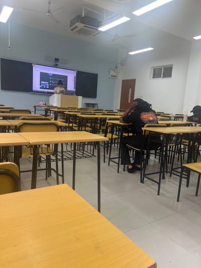 The image depicts a classroom setting with a presentation projected onto a large screen at the front. A person is standing at a lectern, seemingly addressing the class. Several students are seated at desks, with one student in the foreground appearing to be in distress or deep thought, resting their head in their hands. The classroom is equipped with typical school furniture, including desks, chairs, and overhead lighting. The overall atmosphere suggests a lecture or class session is in progress.