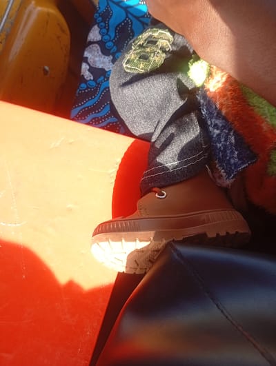 The image shows a close-up view of a person's leg wearing a brown boot and dark jeans. The boot has laces and a thick, ridged sole. The jeans have visible white stitching. The person is sitting in what appears to be a vehicle, indicated by the presence of an orange surface on the left, possibly a seat or panel, and a black, textured surface below the boot, likely another seat. In the background, there is a brightly colored textile with blue and white patterns, and a fuzzy, multicolored fabric in shades of orange, green, and blue. Sunlight casts a strong shadow across the orange surface, suggesting it is daytime and possibly outdoors or near a window. The overall impression is a casual, everyday moment captured in a possibly warm climate, given the bright sunlight. No text is visible in the image.