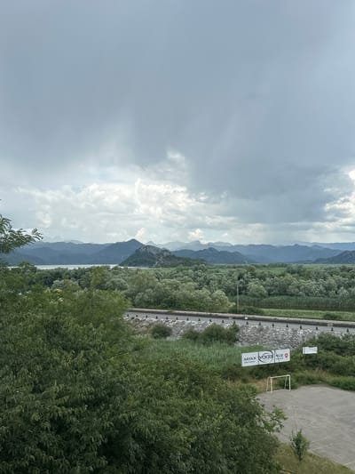 The image is an outdoor, eye-level, landscape photograph of Vranjina, Montenegro, under a cloudy sky. In the foreground, lush green trees dominate the left side of the frame, with their leaves obscuring much of the lower portion. To the right of the trees, a concrete or asphalt surface is visible, with a small, white soccer goalpost situated on it. Behind the goal, a chain-link fence runs horizontally across the frame. Attached to the fence are two white banners with black text; the left banner appears to be reversed text and the right banner has a red circular logo and white text. Beyond the fence, a road runs parallel to the frame, bordered by a stone retaining wall on its near side and dense green vegetation on its far side. In the middle ground, a vast expanse of green trees stretches towards a body of water, likely Skadar Lake, which reflects the muted sky. The background features a range of hazy, blue-hued mountains that fade into the distance, with layered peaks and valleys. The overall atmosphere is subdued, with the overcast sky casting a soft, diffused light over the scene.
