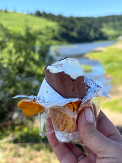 A hand holds a partially unwrapped ice cream bar with a bite taken out of it. The ice cream is coated in chocolate and has a white center. The background is a blurry landscape of green hills and a river under a clear blue sky. The image appears to be taken outdoors in a natural setting, possibly in Spassk, Russia, given the description. The hand has a ring on one finger.