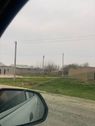 This image depicts a semi-rural street scene in Yettitom, Uzbekistan, observed from the interior of a vehicle. The foreground shows a partial view of a black side mirror reflecting a white car, positioned over a gravel and dirt road surface. A thin horizontal line, likely a scratch, crosses the lower-mid section of the frame. The mid-ground features patches of vibrant green grass and exposed dirt mounds. Several light-colored wooden utility poles with multiple parallel power lines traverse the scene horizontally. On the left, a single-story building with a light grey exterior, a slightly sloped dark roof, two dark doors, and an exterior air conditioning unit is visible. Further back and to the right, several single-story, earth-toned buildings with flat roofs are interspersed among bare deciduous trees and brush. The background consists of a uniform, light grey, overcast sky. No individuals or specific actions are visible within the frame.