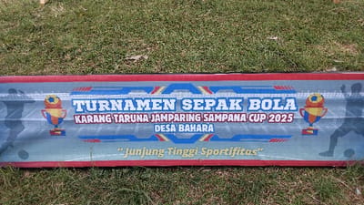 A banner advertising a football tournament is prominently displayed on grassy ground outdoors, likely during daytime. The banner, featuring a blue and red design with white and yellow accents, includes graphic elements such as silhouettes of football players on the left and right, and two trophies topped with footballs.
The visible text on the banner reads:
* "TURNAMEN SEPAK BOLA" (Football Tournament)
* "KARANG TARUNA JAMPARING SAMPANA CUP 2025" (Jampiring Sampana Youth Organization Cup 2025)
* "DESA BAHARA" (Bahara Village)
* "Junjung-Tinggi Sportifitas" (Uphold Sportsmanship)
This indicates an upcoming football tournament organized by the Jampiring Sampana Youth Organization for Bahara Village in 2025, with an emphasis on sportsmanship. The setting in Panjalu, Indonesia, suggests a local community event.