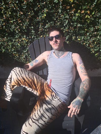 A person with extensive tattoos is reclining on a dark-colored bench outdoors, next to a wall of green foliage. The person is wearing sunglasses, a light gray tank top, and tiger-print pants with a red drawstring. The location appears to be Vicente López, Argentina, based on analysis of foliage and building characteristics. The person is illuminated by sunlight, creating shadows on the bench and clothing.