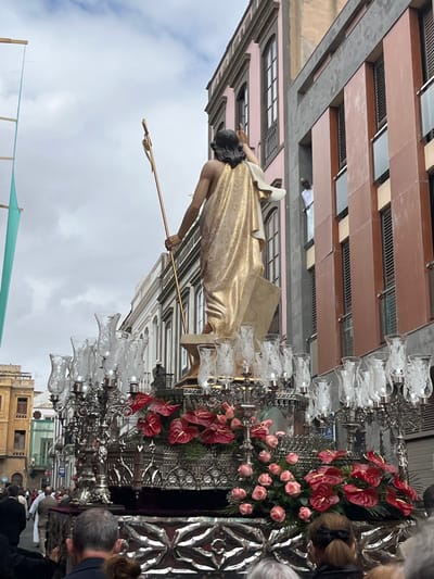 The image captures a religious procession in the city of Las Palmas de Gran Canaria, Spain. The procession is taking place on Calle de Triana, as indicated by the coordinates (latitude: 28.098, longitude: -15.416). The procession features a large, ornate float with a statue of Jesus Christ atop it. The statue is gold-colored and depicts Jesus with his arms raised in prayer. The float is decorated with numerous glass candle holders, red flowers, and white roses. The procession is moving down the street, and the image is taken from a perspective behind the float. Several people can be seen in the background, including a man in white clothing looking out of a window. A crowd of people, some obscured by the float, can be seen in the foreground watching the procession. The image is framed by the buildings on either side of the street. The buildings have multiple stories and feature balconies and windows. The sky is partly cloudy.