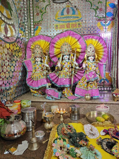 The image depicts an altar in a Hindu temple, likely within a residential setting in Gurgaon, India. The primary subjects are three white marble idols of deities, likely Radha-Krishna or similar figures, dressed in ornate yellow and pink clothing adorned with sequins and gold trim. They wear elaborate crowns and jewelry. The idols are placed on a raised platform, with large, decorative fan-shaped backdrops in yellow and pink behind them. The wall behind the idols is covered in a mosaic of small, reflective tiles in various colors and geometric patterns, with some larger decorative elements.
In the foreground, on a countertop, are various offerings and ritual items. These include several stainless steel containers, some containing what appears to be food items or ghee, and a small brass oil lamp with three lit wicks. There is also a brass bell, a box of matches, and several other containers and packaged goods, one labeled "Milkfood RICH DESI GHEE". A small, ornate brass pot or incense burner is also visible.
A yellow cloth is spread out on the lower part of the countertop, featuring a collection of small, elaborately dressed doll-like figures, likely representing deities or devotees, and various decorative fabrics and ornaments. On a brass plate to the right, two green mangoes are placed as an offering. The lighting appears to be a mix of ambient light and the glow from the oil lamp. The overall impression is one of a devotional space, possibly in a home or a smaller community temple, prepared for worship or a special occasion.