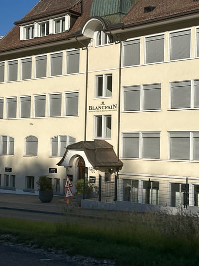The image shows the Blancpain Manufacture in Le Sentier, Switzerland. The building is a multi-story structure with a cream-colored facade and a brown tiled roof. Several windows with gray blinds are visible on each floor. The Blancpain logo and text reading "MANUFACTURE DE HAUTE HORLOGERIE" are prominently displayed on the building. In front of the building, there are several signs, including a traffic sign depicting a worker and a traffic light. A large potted plant is situated near the entrance. The scene is set against a clear blue sky.
