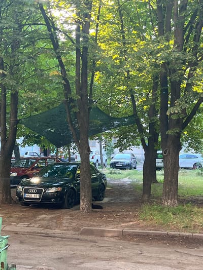 The image depicts a street scene with cars parked between trees. The foreground is dominated by the leafy branches of trees, casting dappled sunlight. Several cars are visible, including a black Audi with the license plate "HNO 101" clearly displayed. Other vehicles, some partially obscured by foliage or other cars, can be seen further down the street. The ground appears to be a mixture of dirt and pavement, with a curb running along the right side of the image. In the background, hints of buildings suggest an urban or suburban setting. The lighting suggests it is daytime, possibly late afternoon due to the warm tones of the sunlight. The overall impression is one of a quiet street in a residential area, likely in Cherkasy, Ukraine. There are no people visible in the frame.