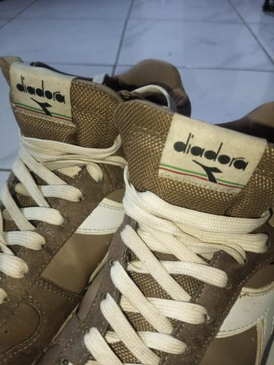 Two well-worn Diadora high-top sneakers are displayed. The shoes feature a brown suede and leather upper, accented by a white stripe on the side. Cream-colored laces crisscross over the tongues. The Diadora logo, surrounded by green, white, and red stripes, is prominently visible on the tongue of each shoe. The backdrop appears to be a tiled floor.