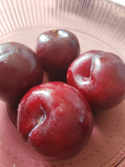 Four dark red plums are arranged in a light pink, ribbed bowl. The plums are glistening with water droplets and are positioned to fill most of the frame. The bowl has a textured surface with a pattern of small circles and lines. The background is a soft blur of pink and white, suggesting diffused lighting. The image appears to be a close-up shot taken from a slightly elevated angle.