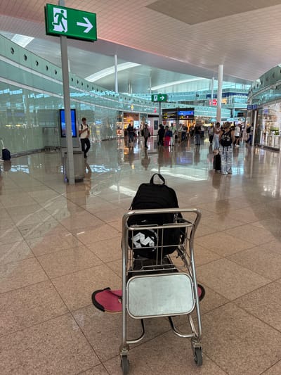 This image depicts the interior of an airport terminal, likely at Josep Tarradellas Barcelona–El Prat Airport in El Prat de Llobregat, Spain, given the visual cues of an international airport environment. In the foreground, a luggage cart is positioned on a polished tiled floor, with a black backpack placed on top. A pink item, possibly a sandal or mat, is visible beneath the cart. The background reveals a busy airport concourse with numerous people walking, some with luggage. Several shops and information signs are visible along the walls, which are lined with reflective glass panels. An illuminated green exit sign with an arrow pointing to the right is prominently displayed on a pole, indicating an emergency exit. The ceiling is made of white modular panels, and large columns support the structure. The overall impression is one of a modern, well-lit, and active airport transportation hub.
