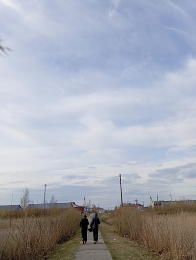 The image depicts a daytime scene in Yurgamysh, Russia. Two individuals are walking along a sidewalk, with buildings and infrastructure visible in the background, including power lines, and structures suggesting residential or industrial areas. A grassy area with dry, brown foliage borders the sidewalk. The sky is overcast with scattered clouds.