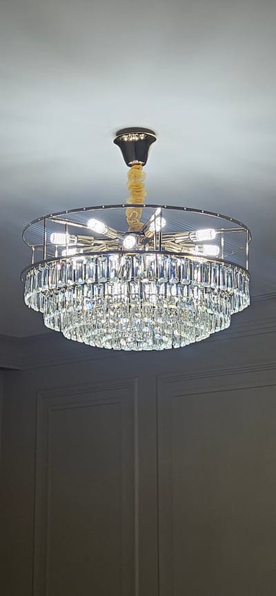 A large, circular chandelier hangs from a white ceiling in a room with paneled walls. The chandelier has multiple tiers of clear, rectangular crystal pendants and is illuminated by several small, white LED bulbs arranged in a circular pattern within its metal frame. A yellow fabric ties the chandelier's cord to its mounting. The paneled walls are a neutral gray color, with vertical lines and rectangular recessed panels. The lighting is bright, reflecting off the crystals. The scene appears to be an indoor setting, possibly a home or a commercial space, in Kaspiysk, Russia. There are no people or animals visible in the image.