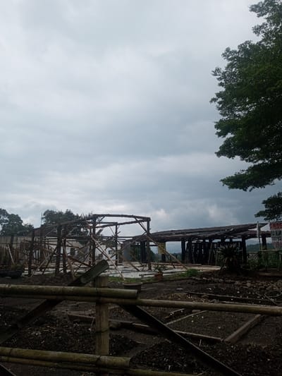 The image displays an outdoor scene in Kandapola, Sri Lanka, under an overcast sky filled with uniform grey clouds. The foreground consists of dark, disturbed earth, appearing as tilled soil or excavated ground, interspersed with small rocks and debris. Multiple bamboo poles are horizontally and diagonally arranged in the immediate foreground, forming a low barrier or an initial framework. In the midground, two distinct, partially constructed or deconstructed open-frame structures are visible. The structure on the left is a skeletal framework of vertical and diagonal wooden or bamboo poles. The structure on the right is a more developed framework with numerous vertical supports and a dark, uneven roof structure, possibly timber or natural material, which appears damaged or incomplete. A small potted plant is situated at the base of the left structure, and a spiky green plant is visible near the right structure. On the far right midground, a partially visible white banner with red text, including fragments like "OMAN," "ALUM," and a telephone number, is affixed to a support. The background reveals dense green tree foliage along the right edge, with distant green hills or mountains extending across the horizon. No individuals are present in the scene, and no discernible actions or interactions are depicted.