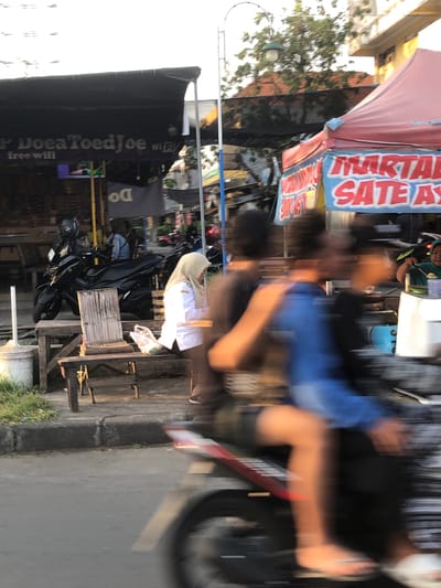 The image captures a bustling street scene, likely in Sumokali, Indonesia, during daylight. The primary focus is on a street vendor's stall, characterized by a red and white striped awning advertising "MARTABAK SATE AYAM" (Chicken Satay Martabak). Several people are present, though most are blurred due to motion, suggesting activity and movement within the scene. One person in the foreground is wearing a blue shirt and appears to be in motion, possibly a customer or the vendor. Another individual in the background is wearing a light-colored head covering and white clothing, seemingly interacting with the stall.

The setting is an outdoor marketplace or street with various objects visible, including motorcycles parked nearby, indicating a common mode of transportation. A prominent black banner with white text reads "DoaeToedJoe free wifi," suggesting a food stall or shop offering internet services. The overall atmosphere appears lively, with the blur of motion hinting at a busy environment. The time of day is likely afternoon or early evening, given the natural lighting. The presence of food and the street vending setup suggests a commercial area where people gather to eat and socialize. The visible text on the awning and banner provides specific details about the products or services offered at the location.