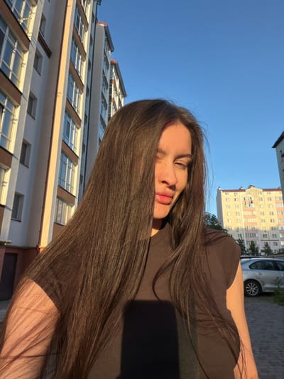 In Vovchynets, Ukraine, a young woman with long, dark brown hair is captured in a selfie outdoors. She is positioned in the foreground, her hair cascading over her shoulders and chest, obscuring parts of her torso. Her face is turned slightly to the right, with her eyes squinted, and her lips are parted. She is wearing a dark, ribbed t-shirt. Sunlight illuminates her face and arm from the right, casting shadows across her body.

Behind her, the scene unfolds with urban elements. To the left, a tall, multi-story residential building with a beige and white facade, accented by brown window frames, dominates the background. Several windows are visible, reflecting the bright blue sky. To the right, another, lighter colored apartment building is visible, with balconies and multiple windows. A silver sedan is parked on a paved pathway in front of the second building. The ground is covered with a textured, light-colored paving stone. The sky is clear and bright blue, indicating fair weather.