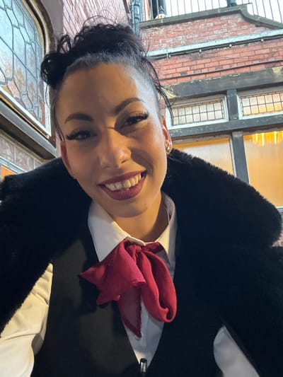 A woman with dark hair, fair skin, and wearing dark lipstick is smiling into the camera. She is wearing a white collared shirt, a dark coat, and a red neck scarf or bow. The background suggests an urban environment with a building constructed of red brick and white-framed windows. Based on the architectural style and the information provided, the photograph may have been taken in Warrington, United Kingdom.