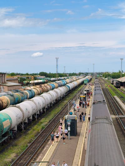 This image depicts a bustling railway station in Buzuluk, Russia. The scene is dominated by long rows of train cars, specifically tank cars used for transporting liquids or gases, parked on multiple tracks. A passenger train is also present, with its roof visible in the foreground, running parallel to the platform.

Numerous people are seen on the platform, walking, talking, and waiting for trains. They are dressed in casual summer attire, suggesting a warm day. The weather appears to be pleasant, with a partly cloudy blue sky. The overall atmosphere is lively and active, indicative of a busy transport hub.

Notable details include the industrial setting with several buildings and structures in the background, along with power lines and transmission towers, typical of a rail yard. There are also trees and greenery scattered around, softening the industrial landscape. A small kiosk or shelter is visible on the platform, and some signage is present, though the text is not clearly legible. The time of day appears to be daytime, likely late morning or afternoon, given the lighting conditions. The presence of people suggests a moment of transit or waiting, with a general sense of movement and activity.