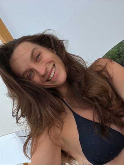 A woman with long, wavy brown hair is captured in a close-up, low-angle selfie. She is wearing a dark blue bikini top with subtle embellishments. Her eyes are looking towards the camera, and she has a wide, joyful smile, showing her teeth. The background is blurred, with a hint of greenery suggesting an outdoor setting, possibly near Drobnići, Montenegro. A large white structure, possibly an umbrella or canopy, is visible in the upper portion of the image, with a wooden support beam on the left. The lighting suggests it is daytime.