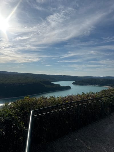 An aerial shot captures a scenic view of a wide, winding body of water, likely a lake or reservoir, surrounded by lush, tree-covered hills. The sun shines brightly in the upper left quadrant of the frame, casting a lens flare and illuminating the wispy clouds that streak across a clear blue sky. In the foreground, a dark metal railing borders a dense green bush line, which overlooks the expansive waterway. The water itself is a tranquil turquoise, reflecting the daylight and the surrounding foliage. Boats can be spotted dotting the surface of the water, and the terrain on the opposite shore appears more populated with buildings visible amidst the trees. The overall impression is one of natural beauty and serene leisure, potentially a viewpoint above the Lac de Vouglans near Moirans-en-Montagne, France, a popular tourist destination known for its vast reservoir and surrounding natural landscapes.