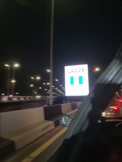 An aerial view shows the busy streets of Ikoyi, Nigeria, at night. The city is illuminated by numerous streetlights and a large, brightly lit digital billboard displaying the word "LAGOS" in large white letters, with two teal-colored rectangles below. The billboard is positioned above a highway, with traffic visible in the distance. The image is captured from within a moving vehicle, indicated by the presence of a side mirror and the blurred motion of the car's interior. The highway has concrete barriers along its sides, and the overall scene suggests a vibrant urban environment.