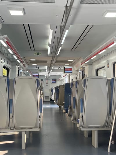 The image shows the interior of a train carriage, likely at the train station in Figueres, Spain, given the destination displayed on the screens. The carriage is empty, with rows of seats on either side of a central aisle. The seats are upholstered in a light gray fabric with blue armrests and are mounted on white metal legs. Overhead, fluorescent lights illuminate the space, and there are digital screens displaying the destination "DESTINO RII PORTBOU" in red text on a white background. Emergency exit signs with green figures are also visible. The walls and ceiling are painted white, with some exposed metal fixtures and red safety railings. The floor is a dark gray material, reflecting the overhead lights. Natural light enters from windows on the sides of the carriage, casting shadows on the floor. The overall impression is one of cleanliness and modernity.